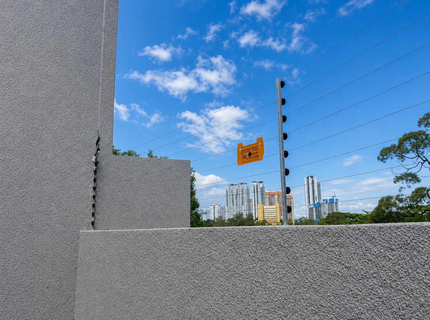 Electric fence posts, parallel wires and warning signage along a textured boundary wall.